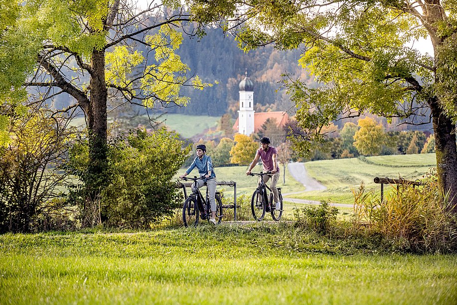 Zwei Radler auf dem Radweg zwischen Bäumen, dahinter eine Kirche Zwei Radler auf dem Radweg zwischen Bäumen, dahinter eine Kirche