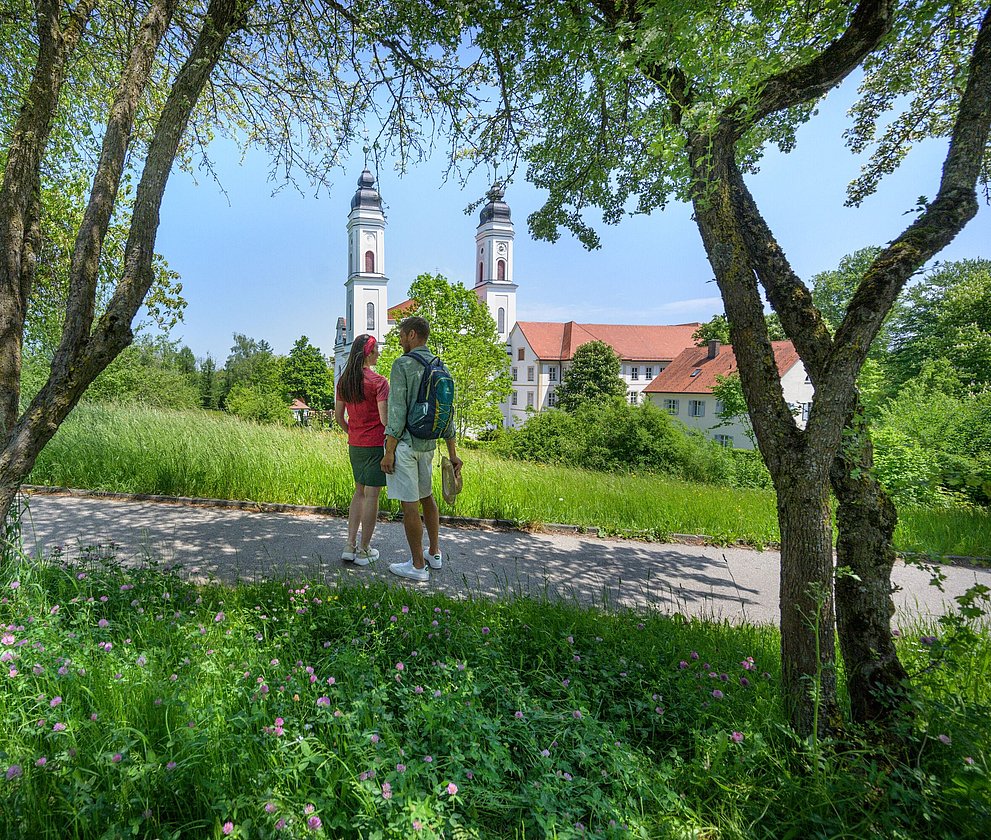 Zwei Personen gehen im Grünen spazieren, im Hintergrund eine Kirche