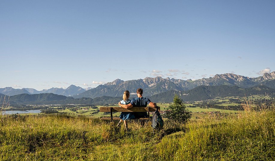 Zwei Wanderer sitzen auf einer Bank, hinter ihnen das Alpenpanorama