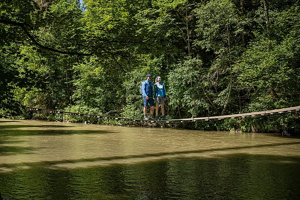 Zwei Personen auf einem Hängesteg über einem Fluss Zwei Personen auf einem Hängesteg über einem Fluss